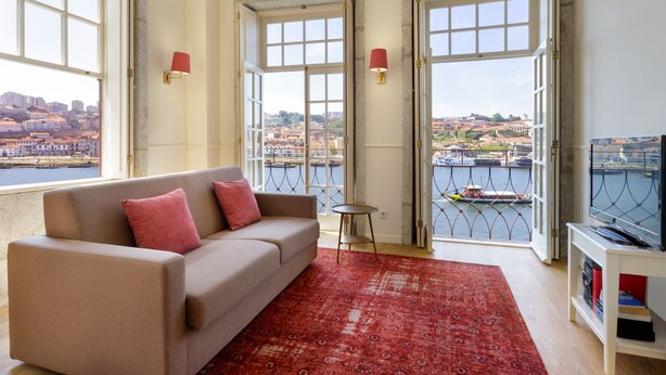 Un salón con sofá, alfombra roja y vistas a un paisaje urbano a través de amplios ventanales.