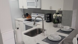 Een moderne keuken met witte kastjes, een magnetron, oven en spoelbak. Er staan twee eettafels met stoelen, en een vaas met bloemen op het aanrecht.