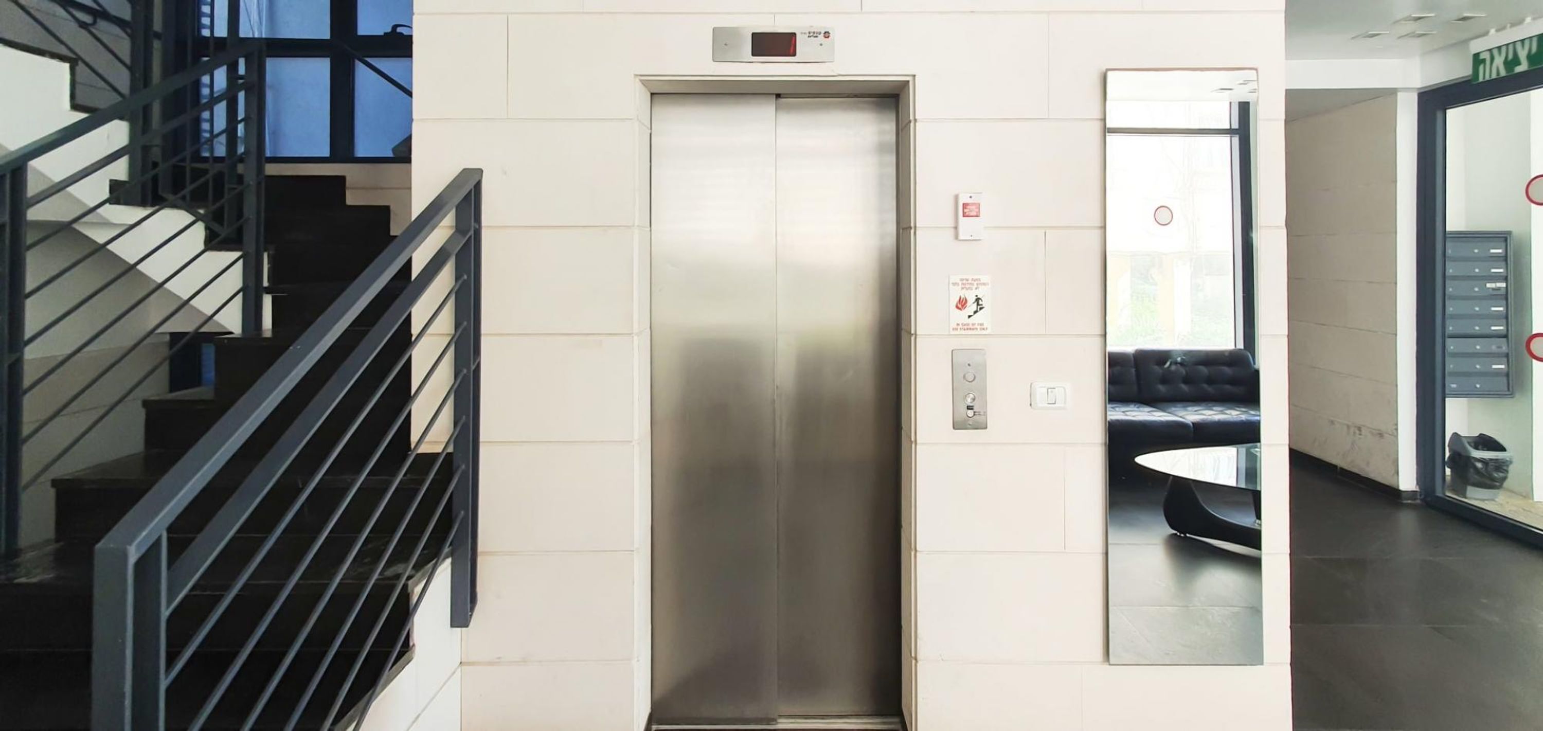 Un ascenseur dans un immeuble moderne aux finitions métalliques, un panneau indiquant le système d’alarme incendie et une porte en verre qui reflète un espace détente avec des sièges.