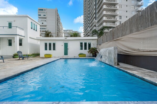 A swimming pool with a waterfall feature, surrounded by white buildings with balconies and a fence.
