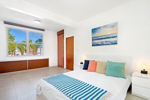 A bedroom with a large window, a bed with multiple pillows, a nightstand with a lamp, and a painting of a beach scene. - Puerto Rico Ocean Park Loft Vacation Rental With Parking Otium Rentals (San Juan)