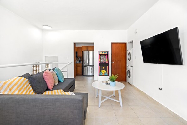 A modern living room with a grey sofa, colorful cushions, a small white table, and a flat-screen TV mounted on the wall. - Puerto Rico Ocean Park Loft Vacation Rental With Parking Otium Rentals (San Juan)