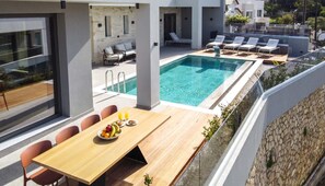 Un área de piscina moderna al aire libre con mesa y sillas de madera, un espacio de descanso con tumbonas y un muro de piedra.