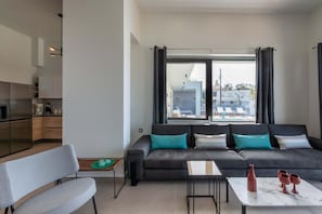 A modern living room with a sofa, a dining table, and a kitchen area visible through an open door. - Lusol Seafront Villa I-free Heated Pool 100m Sea (Chania)