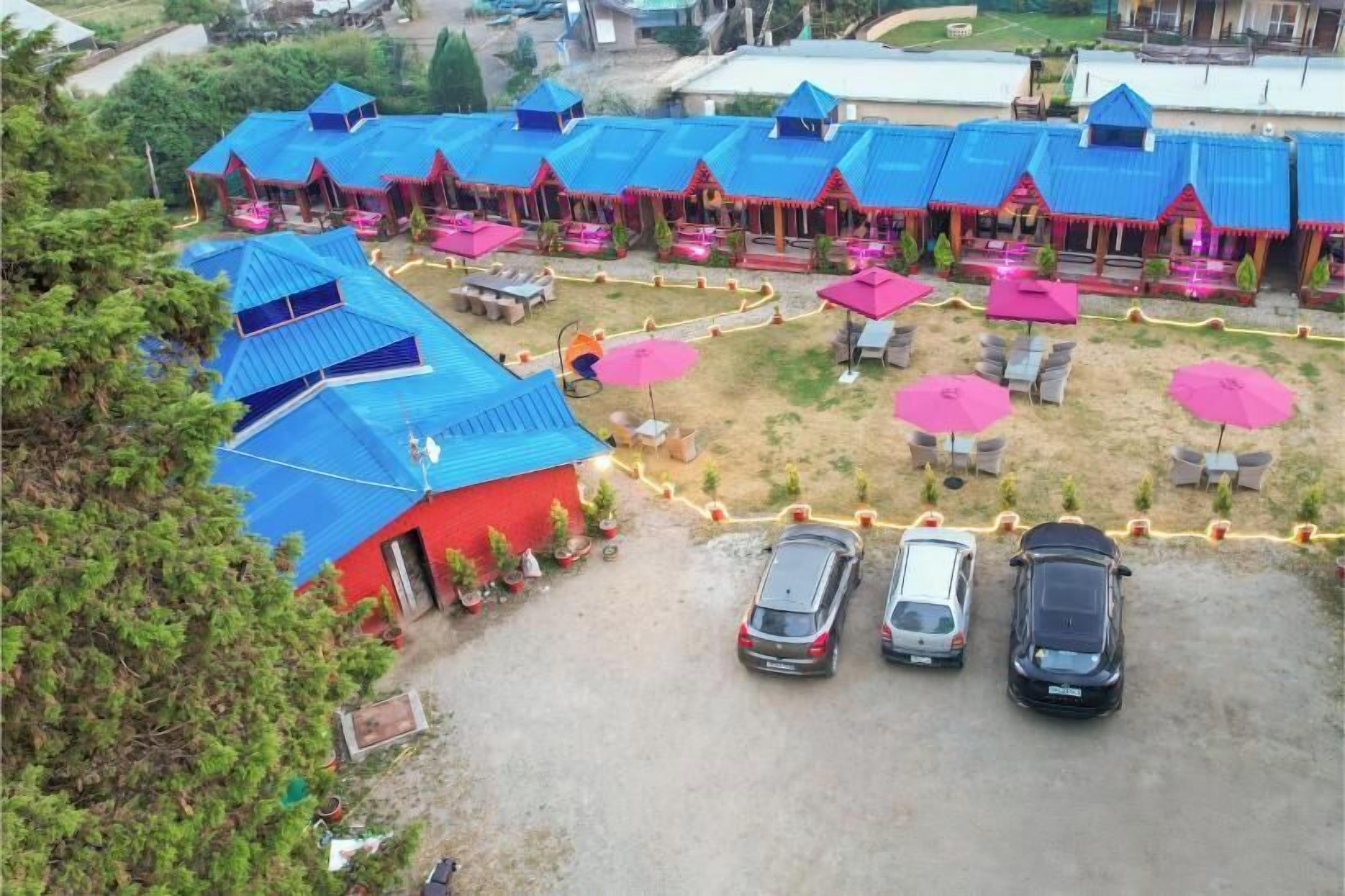 Un complejo turístico con varias construcciones de techo azul, zonas de estar al aire libre con sombrillas rosadas y un aparcamiento con varios coches.