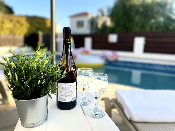 A bottle of wine, two glasses, and a potted plant on a table by a pool.