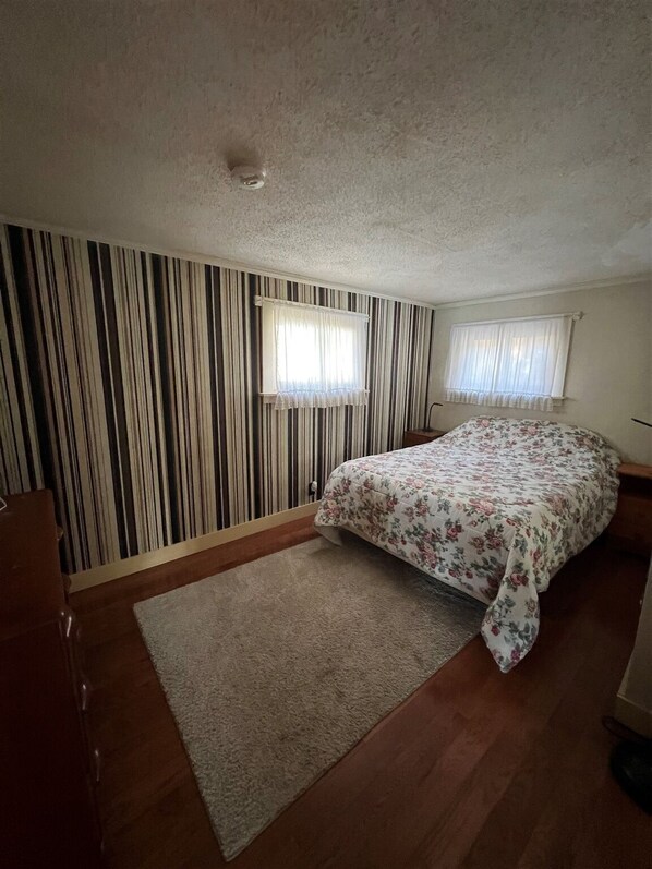 A bedroom with striped wallpaper, a floral bedspread, a wooden dresser, and a small window with curtains.