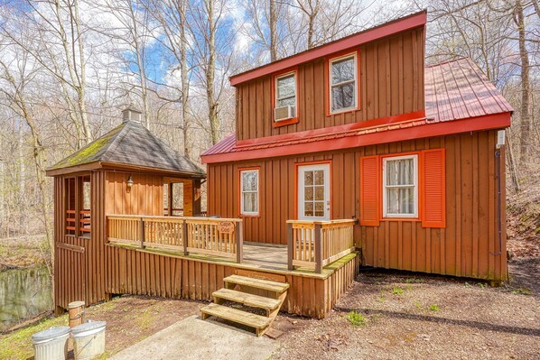 A wooden cabin with a red roof, a small porch, and a dock extending into a body of water.