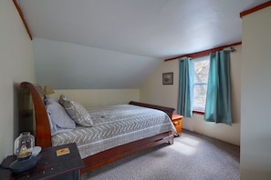Een slaapkamer met een houten bed, een nachtkastje, een lamp, een raam met gordijnen en een klein schilderijtje aan de muur.