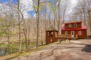 A cabin with a red roof and wooden exterior, situated by a lake surrounded by trees.