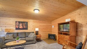 Interior acogedor de una cabaña de madera con sofá, mesa de centro de madera, televisor, chimenea y mecedora.