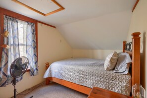 A bedroom with a sloped ceiling, a bed with a patterned bedspread, a standing fan, and a window with patterned curtains.