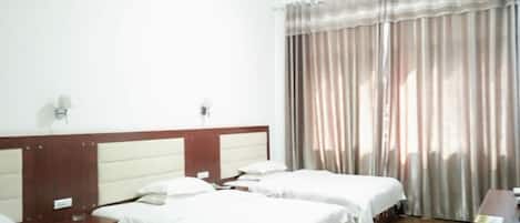 Habitación de hotel con tres camas individuales, cabecera de madera, escritorio y un ventanal con cortinas.
