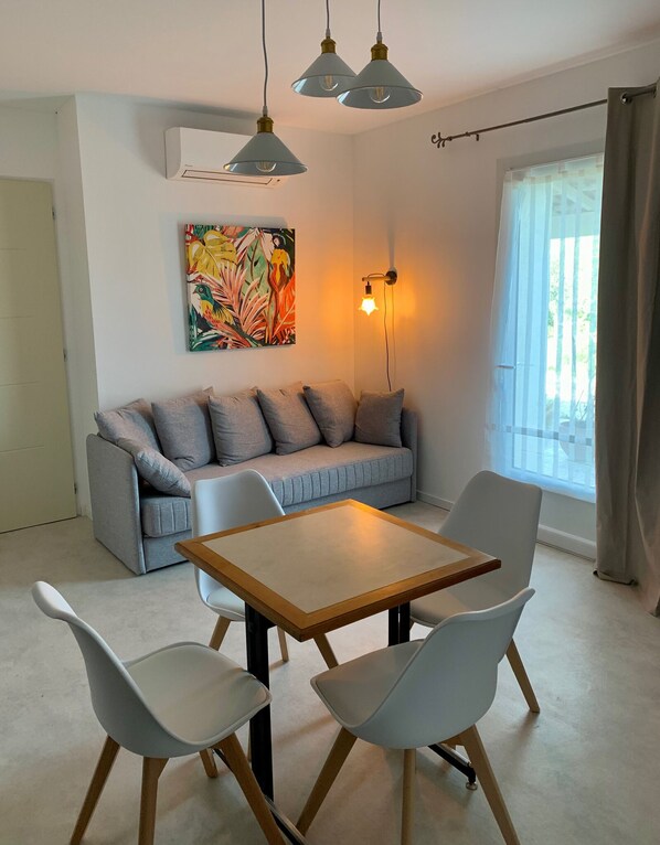 Un salon moderne avec un canapé gris, une table en bois et deux chaises blanches. Un tableau abstrait aux couleurs vives orne le mur, un climatiseur est fixé au plafond et une fenêtre est agrémentée de rideaux.