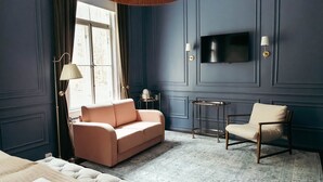 A room with a sofa, a chair, a small table, a TV mounted on the wall, and a window with curtains. - Billnäsin ruukki (Raseborg)