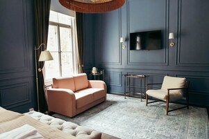 Habitación con sofá, una silla, una mesita, televisor montado en la pared y ventana con cortinas.