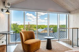 Un balcón moderno con una silla de cuero, una mesita redonda y vistas a un área boscosa con edificios al fondo.