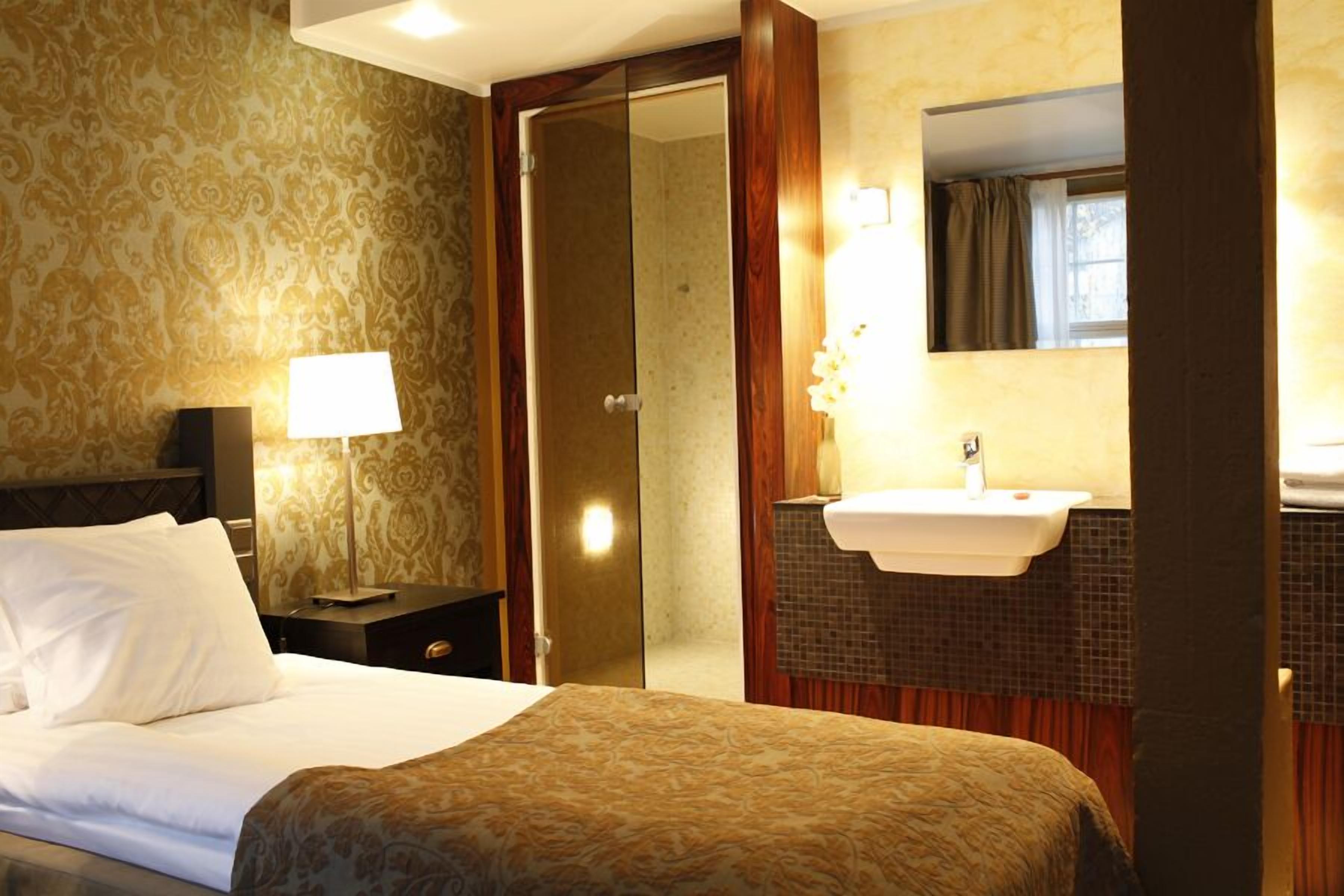 A hotel room with a bed, a nightstand with a lamp, a bathroom with a sink and mirror, and patterned wallpaper.