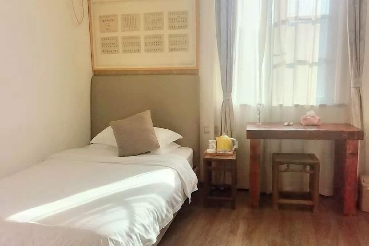 A neatly made bed with a pillow, a small wooden table with a yellow kettle, and a framed chart on the wall.