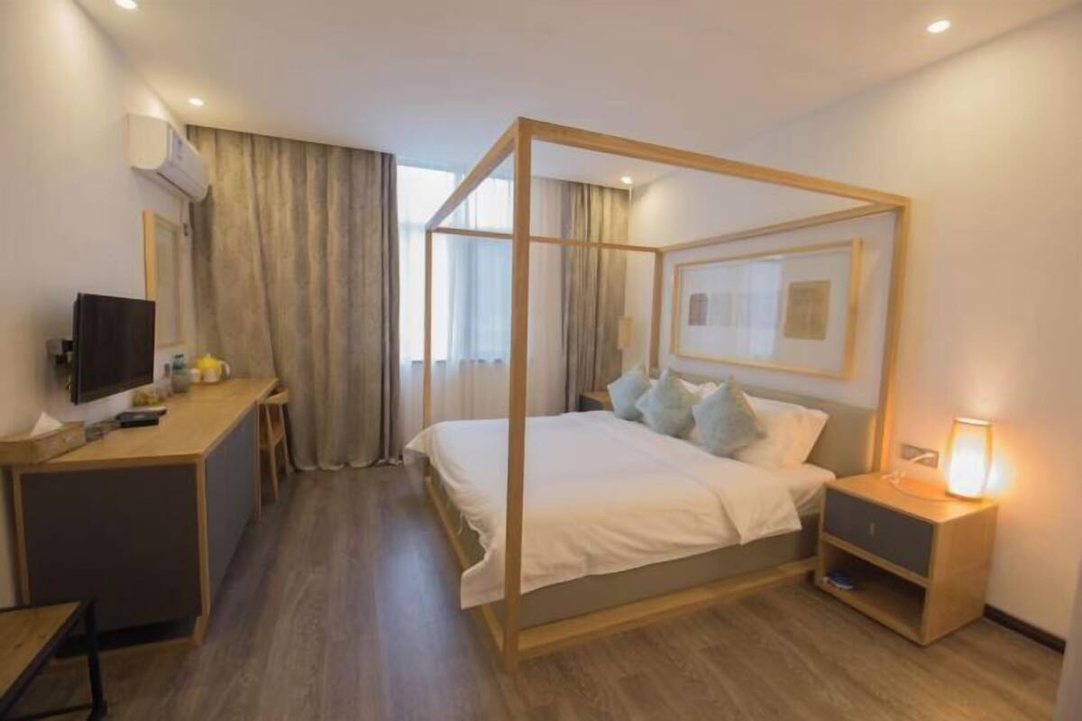 A modern hotel room with a four-poster bed, a desk with a flat-screen TV, and a bedside table with a lamp.