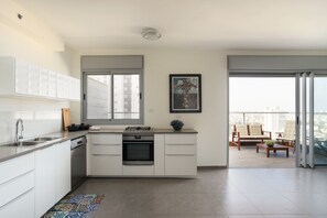 A modern kitchen with white cabinets, a gas stove, and a dishwasher. There is a large window and a balcony with outdoor furniture.