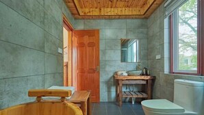 Un baño con bañera de madera, inodoro, lavamanos con espejo y ventana que deja entrar luz natural.