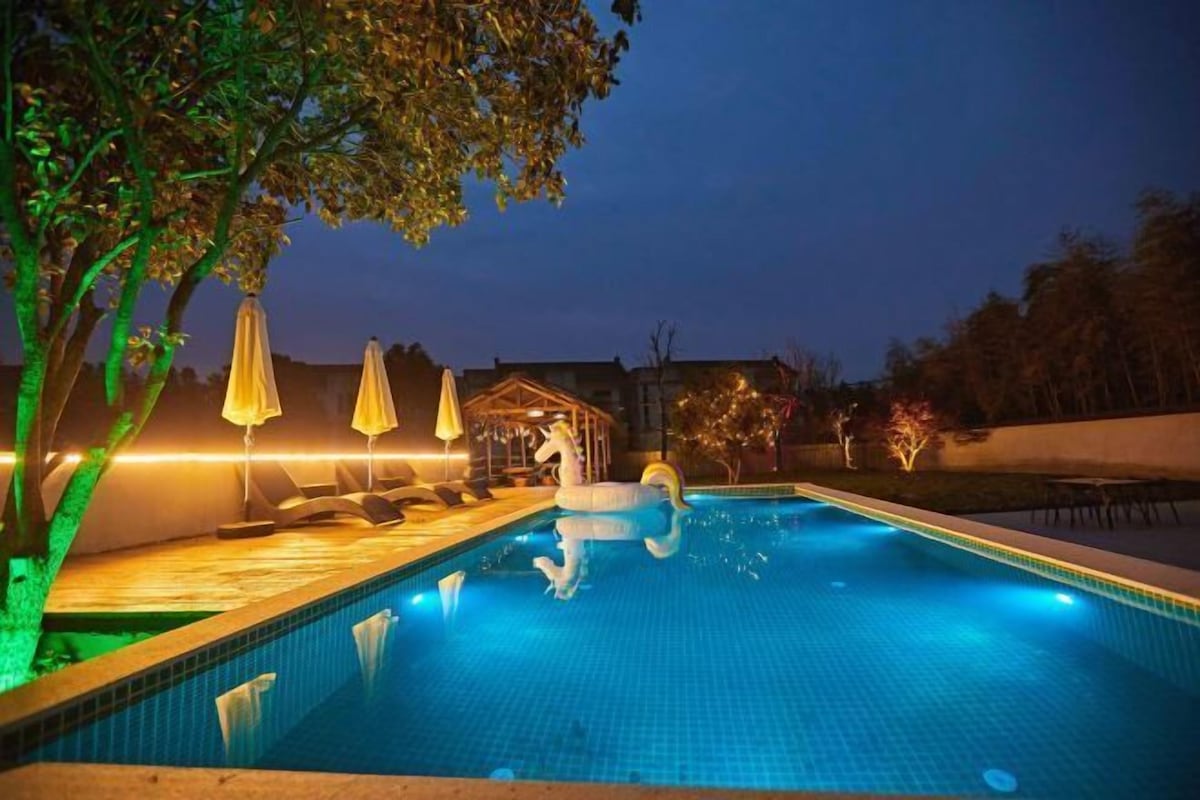 Una piscina amb un flotsam de cigne, envoltada d'arbres i mobiliari exterior amb ombrelles.