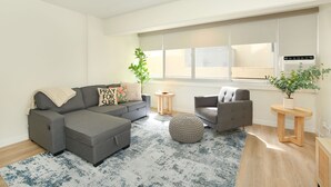 Ein modernes Wohnzimmer mit einem grauen Ecksofa, einem runden Hocker, einem grauen Sessel, einem kleinen Holztisch und einem Blumentopf.