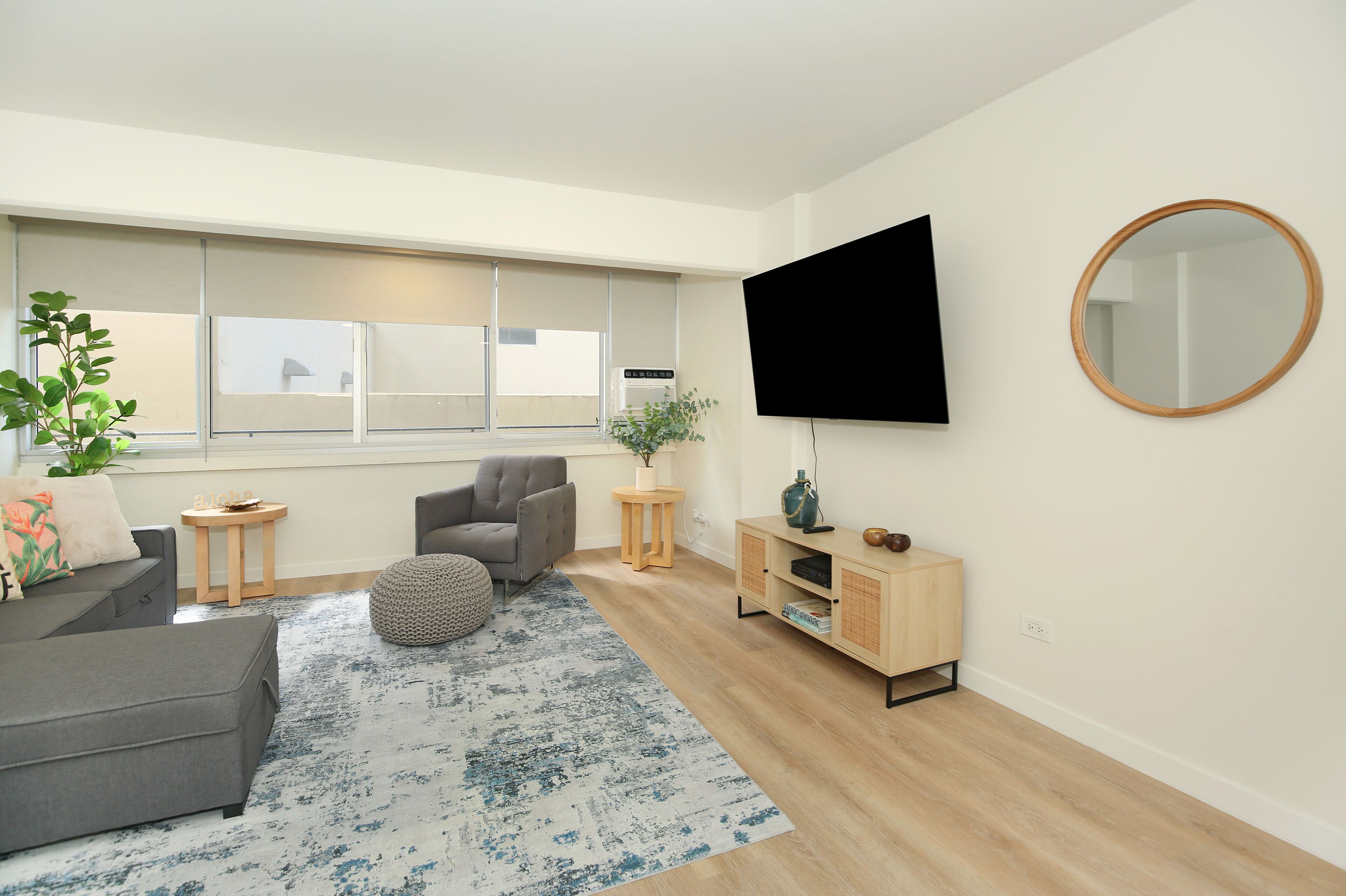 A modern living room with a grey sofa, a round ottoman, a wooden coffee table, a TV mounted on the wall, and a large window with blinds.