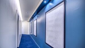 A hallway with blue carpet, white walls, and large windows with blinds. - Sheva Boutique Hotel (Milan)