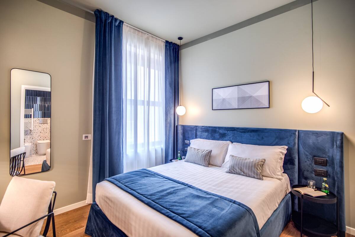 A modern hotel room with a large bed, a side table, a chair, a mirror, and a window with curtains.