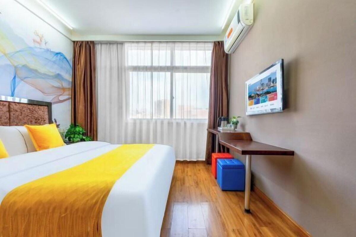 Une chambre d’hôtel comprenant un lit, un bureau, une chaise, une fenêtre avec des rideaux, un téléviseur fixé au mur et un tableau.