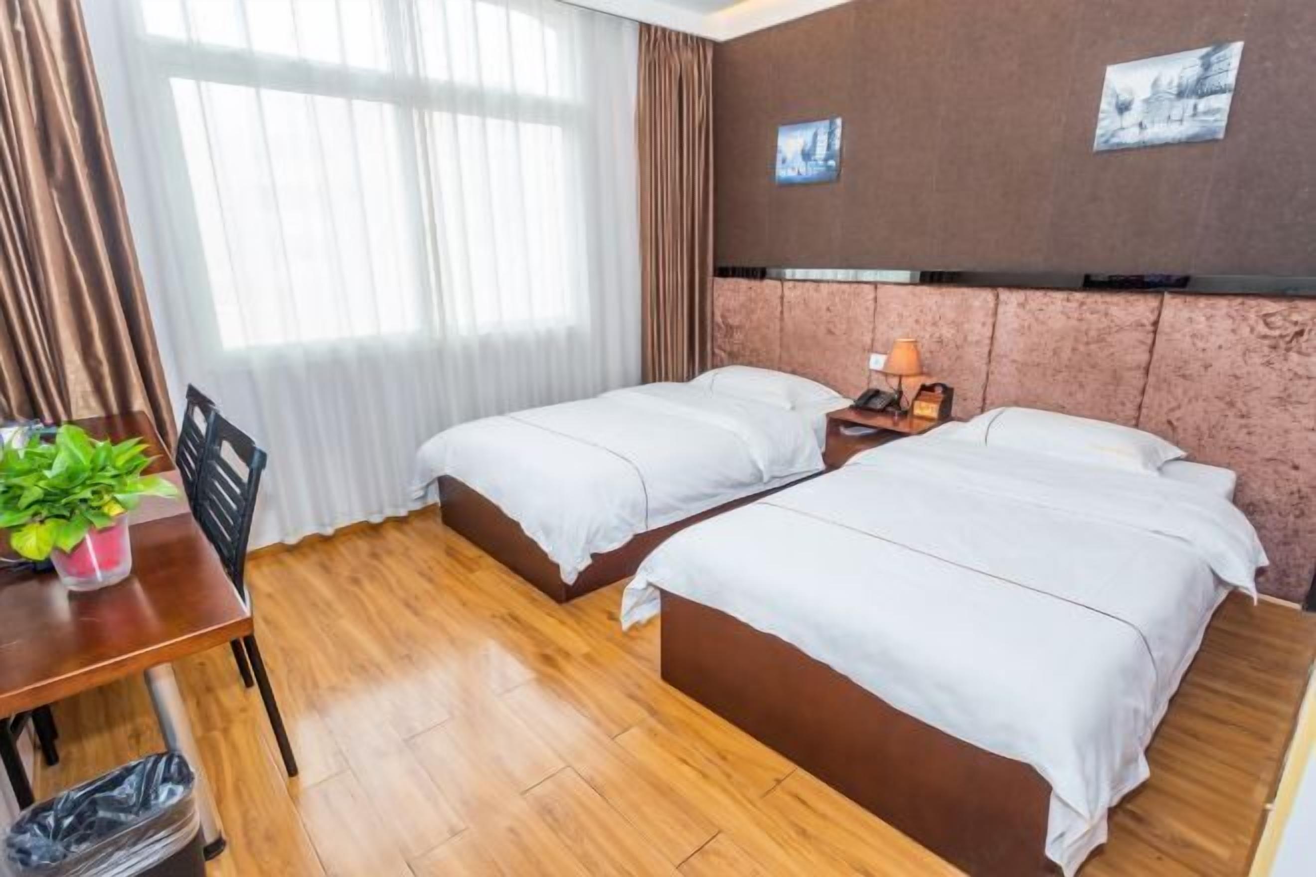 A hotel room with two single beds, a wooden floor, a desk with a chair, a small table with a plant, and a trash bin.
