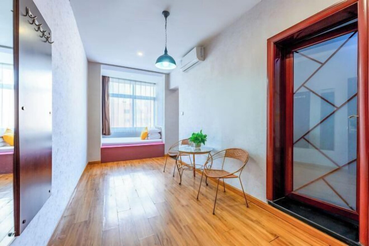 Une pièce avec un plancher en bois, une table ronde entourée de chaises, une fenêtre avec des stores et une porte à cadre rouge.