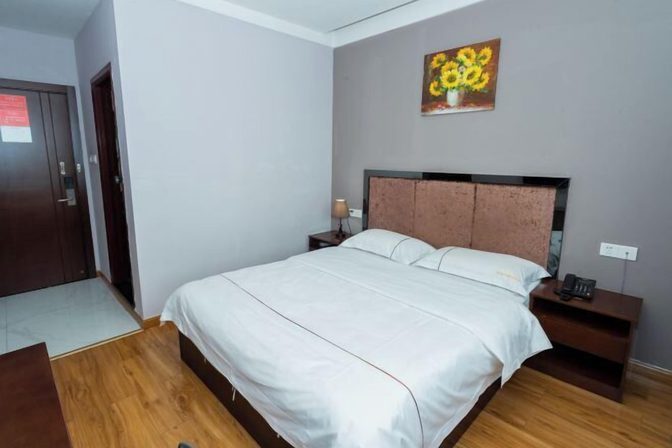 A bedroom with a bed, wooden floor, a nightstand, a painting, and a door.