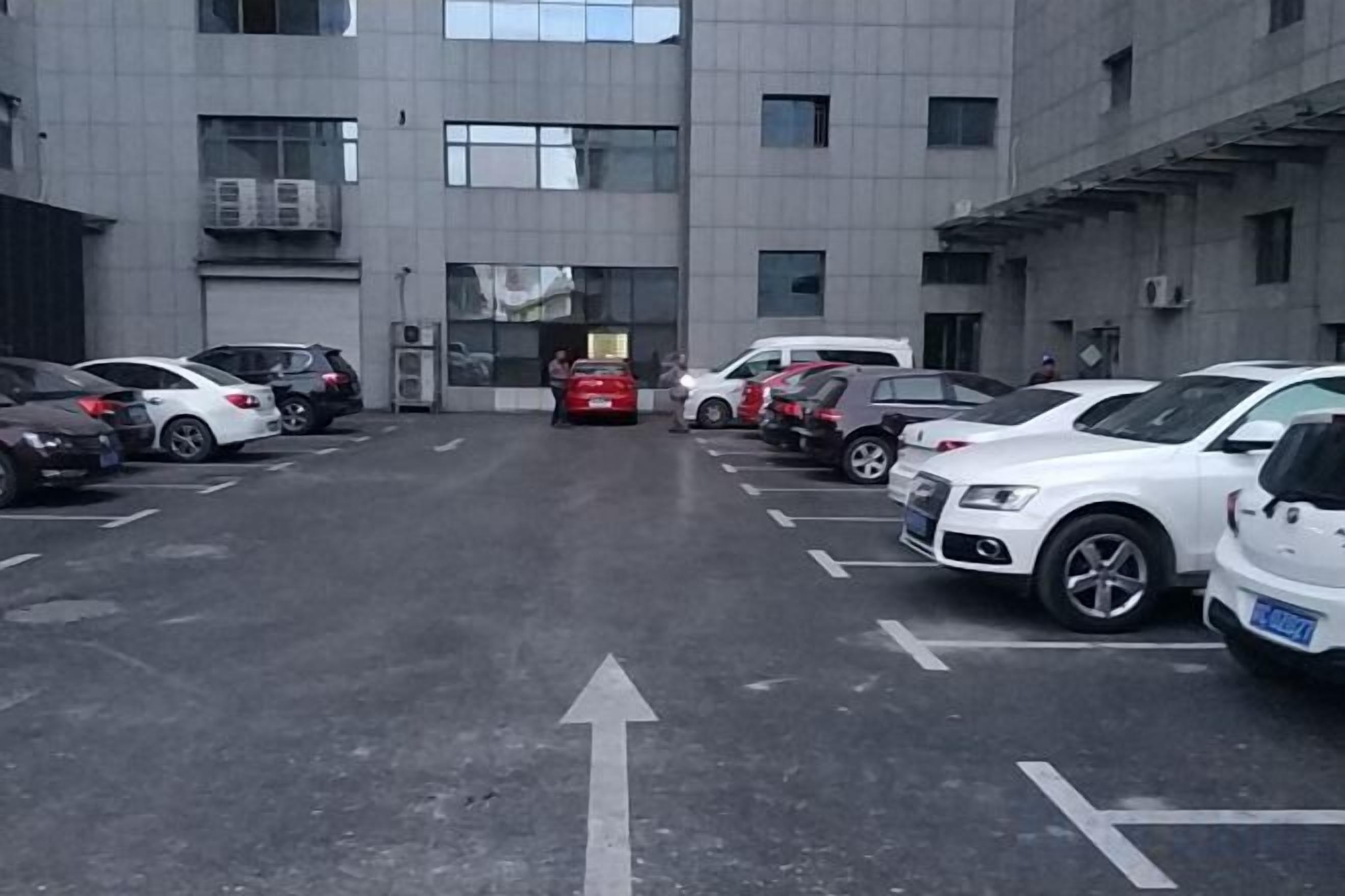 A parking lot with multiple cars parked in designated spaces, a building with a flat facade, and a clear sky.