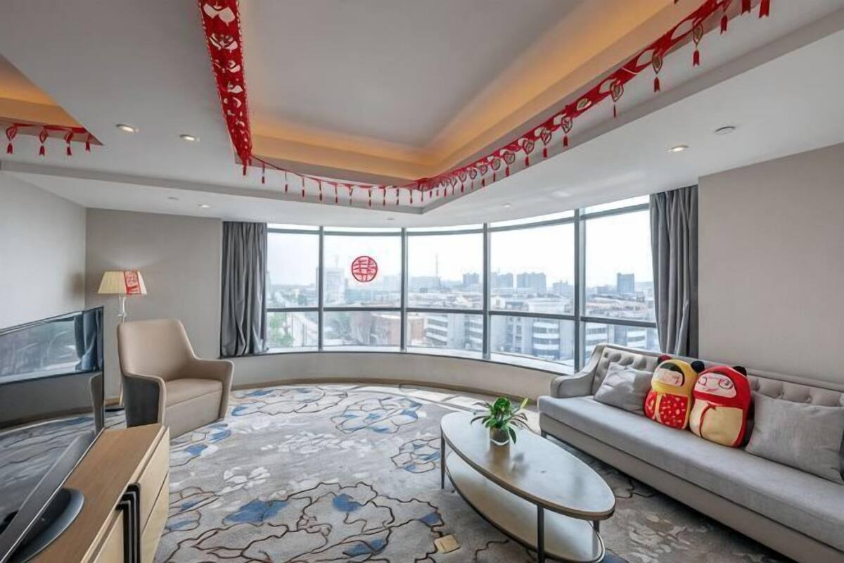 A modern hotel room with a curved sofa, a flat-screen TV, a coffee table, and a large window offering a city view.