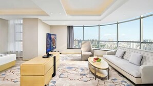 Habitación de hotel moderna con un ventanal grande, un televisor de pantalla plana, un sofá, una silla y una pequeña mesa con fruta.