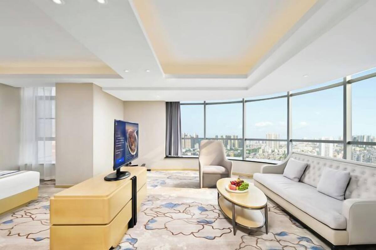 A modern hotel room with a large window, a flat-screen TV, a sofa, a chair, and a small table with fruit.