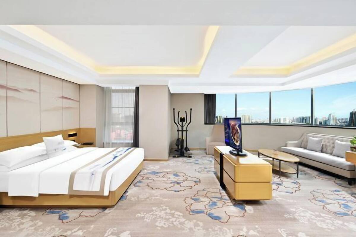 A modern hotel room with a large bed, a sofa, a TV, and a city view.
