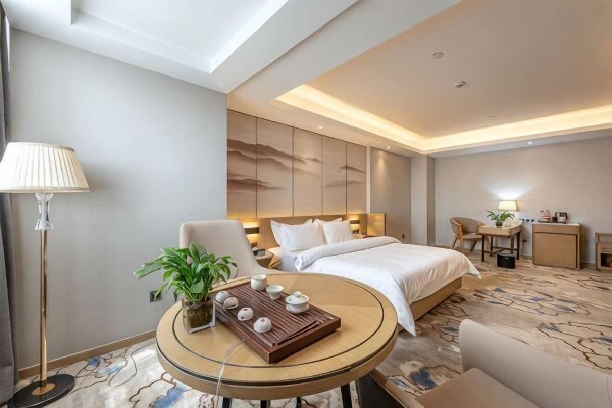 A hotel room with a bed, a desk, a chair, a lamp, a table with a tray of teacups, and a potted plant.