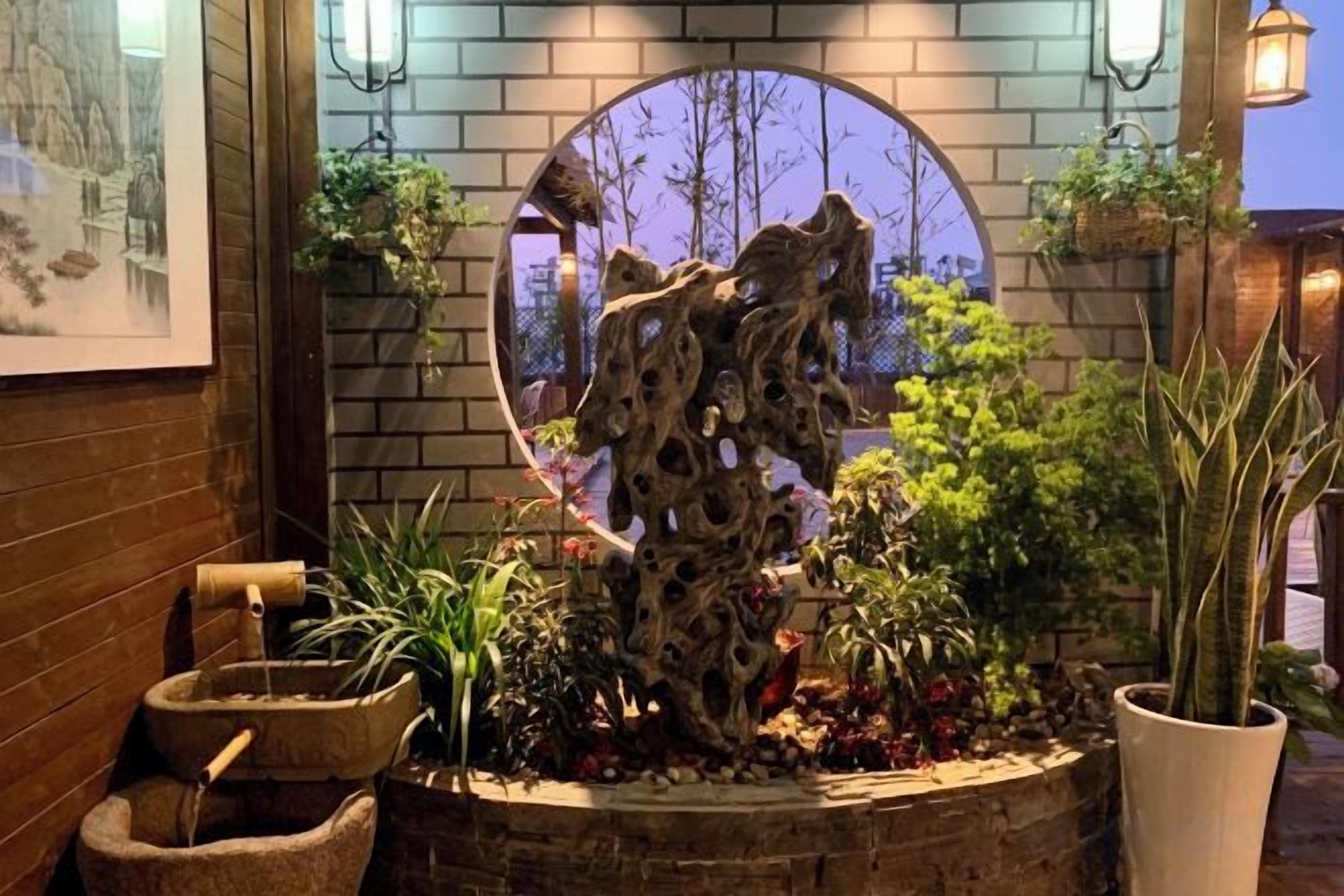 Una jardinera de piedra con un corte circular, rodeada de plantas diversas y una escultura rústica.