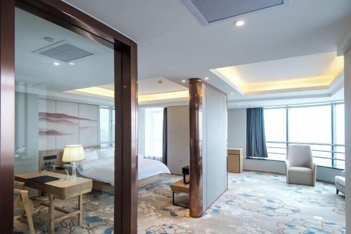 A hotel room with a bed, a desk, a chair, a lamp, and a large window.