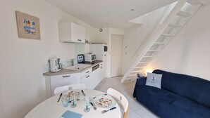 Een kleine keuken met witte kastjes, een magnetron en een koffiezetapparaat. Een eettafel met borden en bestek is gedekt voor twee personen. In de woonruimte staat een blauwe bank en een klein nachtkastje met een lamp.