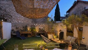 An outdoor dining area with a stone wall backdrop, a wooden table set for a meal, and a hanging light fixture. - Maison Simone, cosy and private garden - Centre Arles - 2 bedrooms. (Arles)