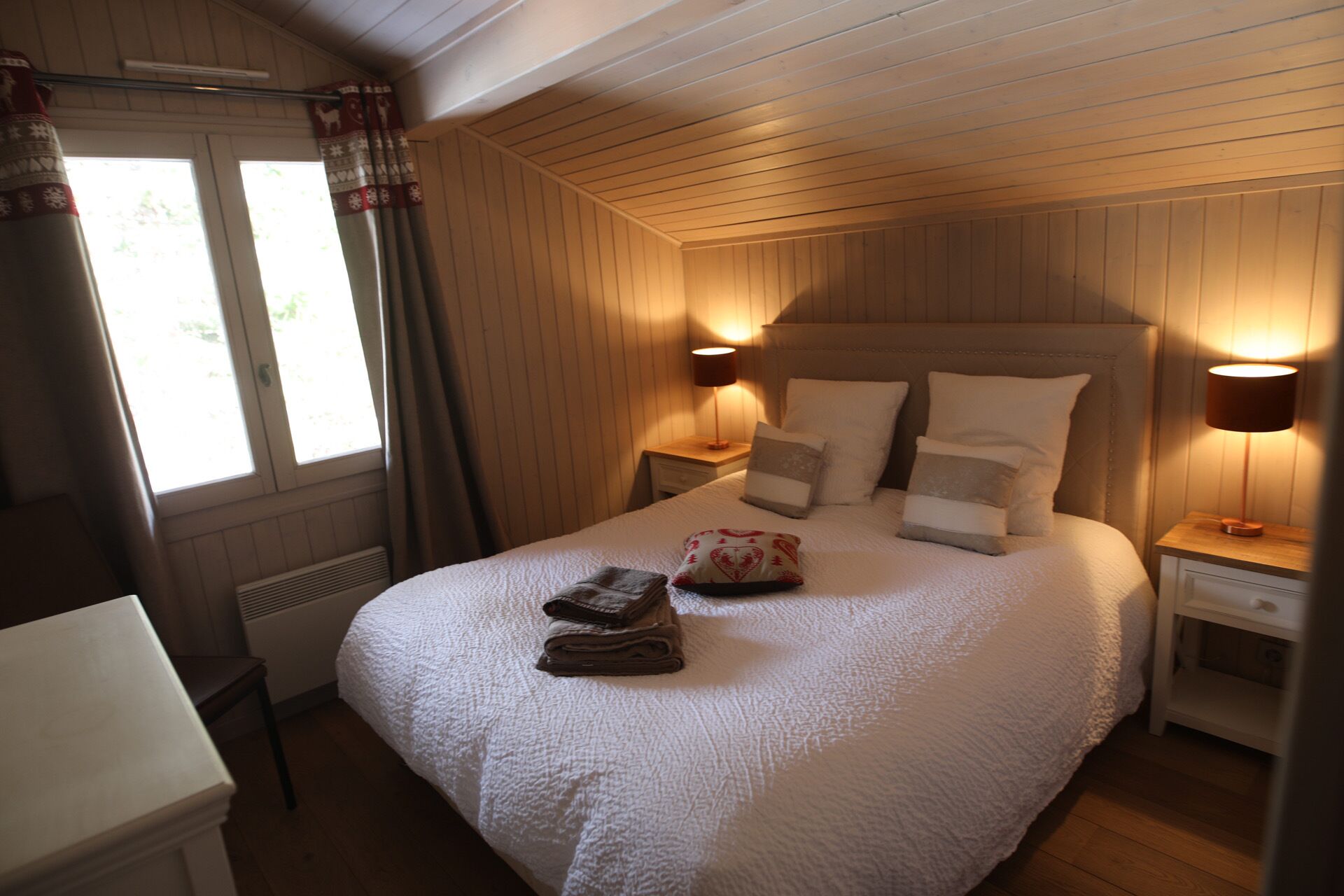 A neatly made bed with a white quilt, two bedside tables with lamps, and a window with curtains.