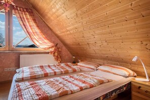 Une chambre mansardée chaleureuse avec des lambris en bois au plafond, une grande fenêtre avec des rideaux, deux lits avec une literie à rayures et une table de chevet avec une lampe.