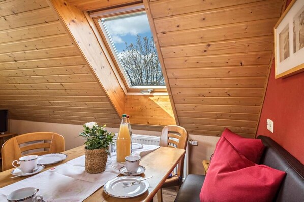 Un coin repas avec une table en bois, des chaises et un canapé rouge. Une fenêtre offre une vue sur les arbres.