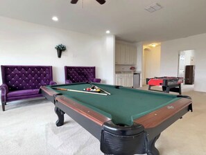 Una mesa de billar con superficie de fieltro verde, rodeada por un sofá morado y un sillón capitonado.