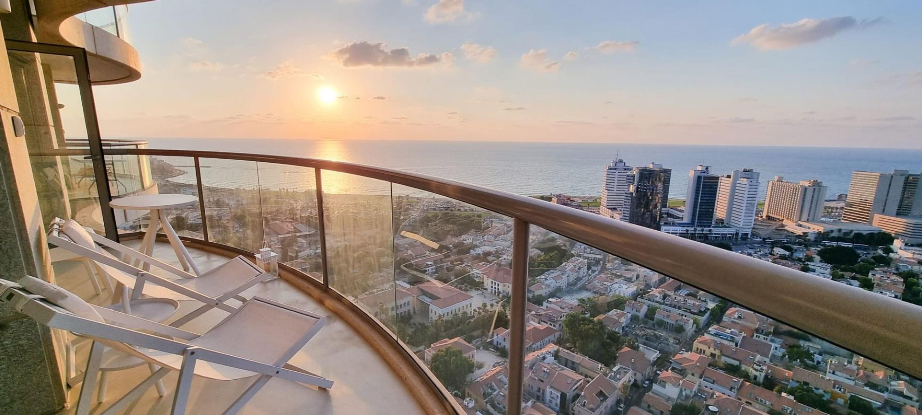 A balcony with a curved glass railing overlooking a cityscape and the sea during sunset.